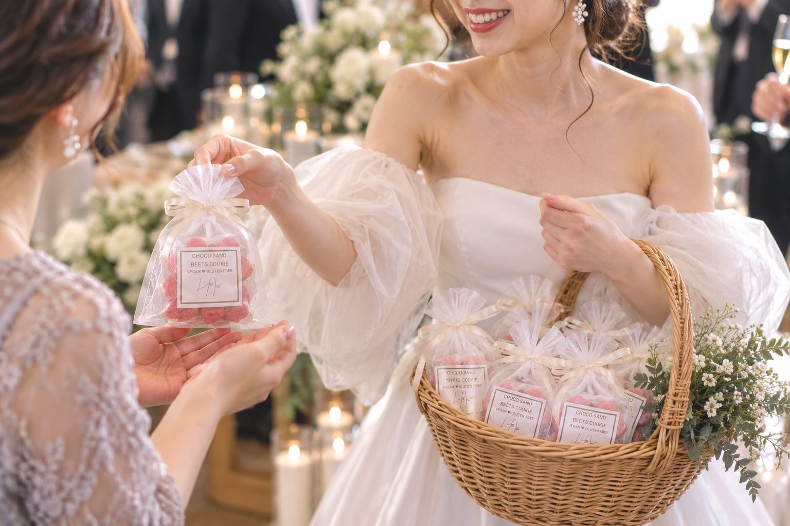 白いドレスの花嫁が、籠に入れたギフトから1つを取り、ゲストに手渡している。背景にキャンドルと白い花