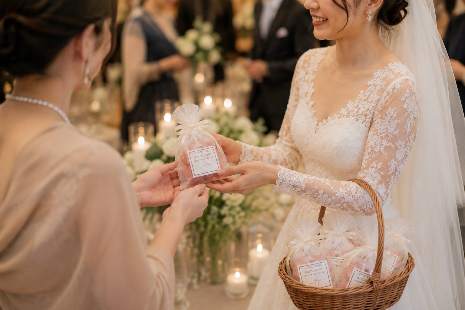 レースのドレスとベールの花嫁が、透明の袋に入れたピンクの焼き菓子をゲストに手渡している。腕には同じ袋を入れた籠