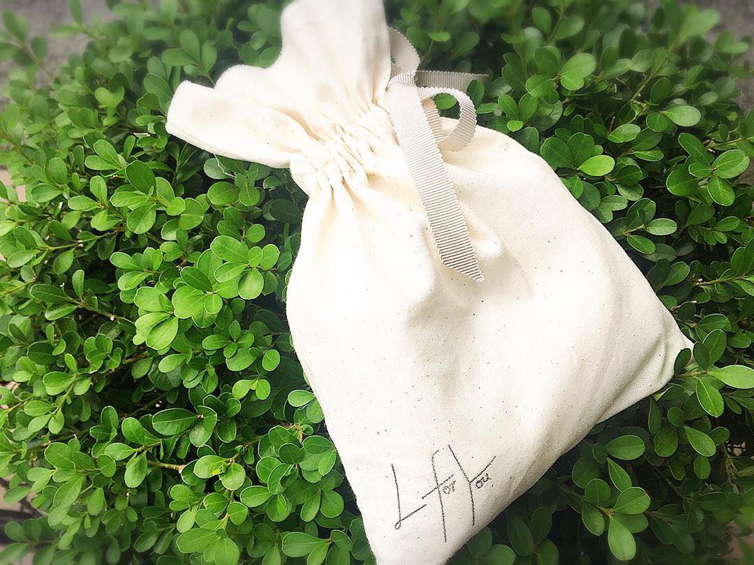 L for You のロゴを刻印した生成りの布の巾着。グレーのリボンで口を絞り、緑の植え込みの上に置いてある